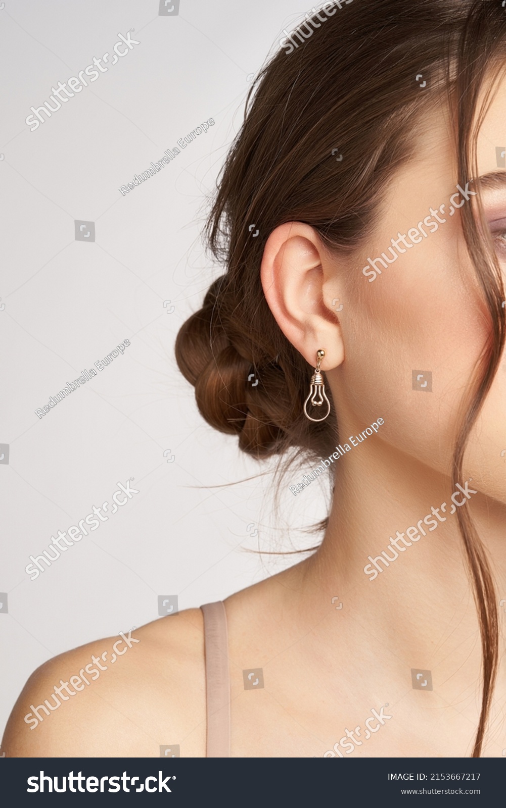 Cropped side portrait of a brown-haired lady in golden round stud earrings with a pendant made as an incandescent light bulb. The girl is posing on the gray background. #2153667217