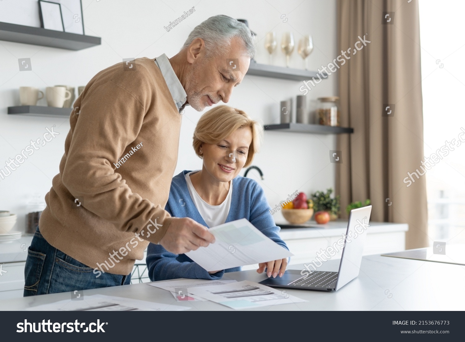Middle aged couple checking finance account together. Wife and husband fill tax form, holding current expense document in hands, sitting on kitchen, paying utility bill online #2153676773