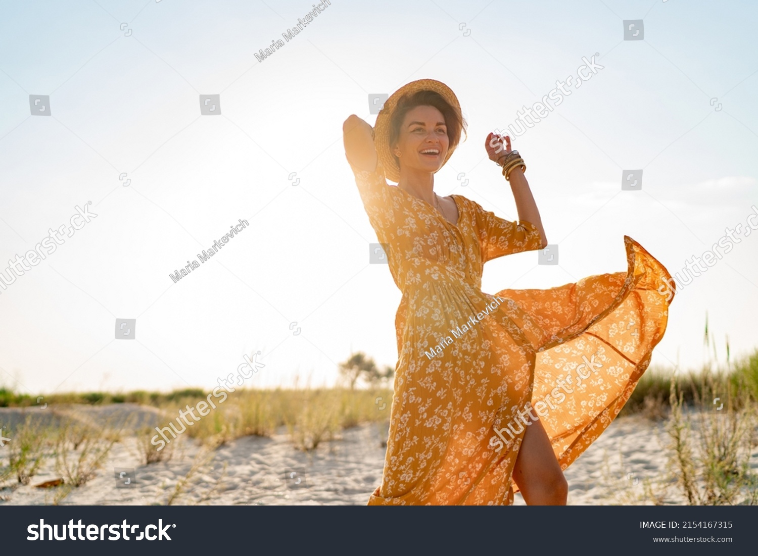 stylish attractive slim smiling woman on beach in summer style fashion trend outfit carefree and happy, feeling freedom, wearing yellow printed dress boho style chic and straw hat #2154167315