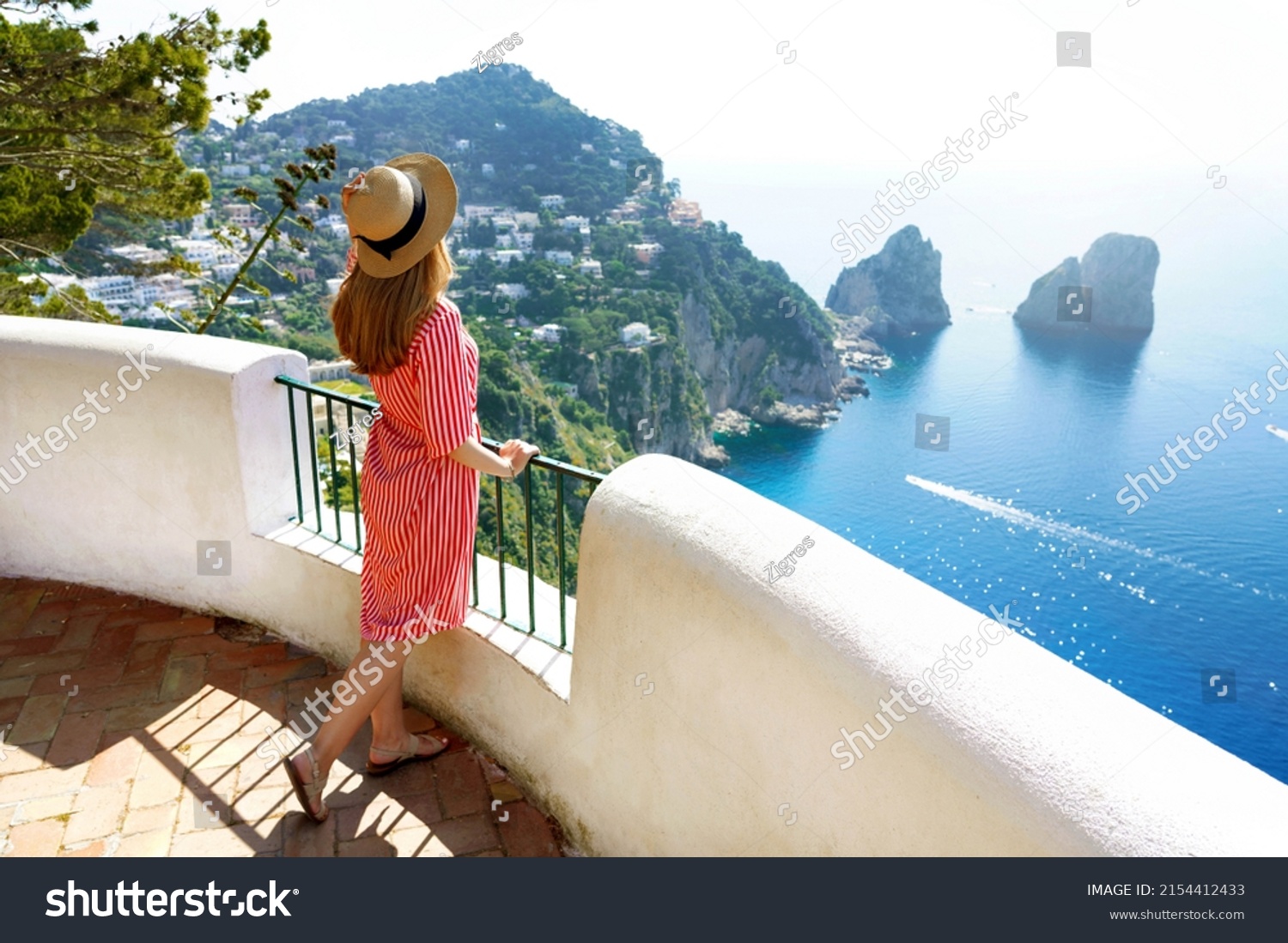 Holidays on Capri Island. Back view of beautiful fashion girl enjoying view of the Faraglioni stacks sea in Capri. Summer vacation in Italy. #2154412433