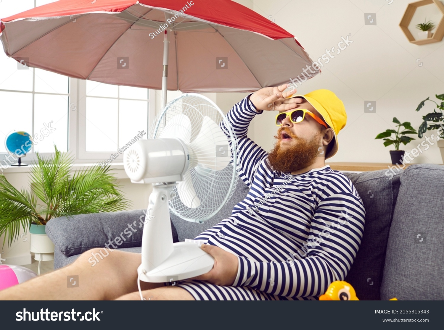 Funny sweaty chubby bearded man in swimsuit sitting at home, suffering from crazy summer heat, wiping sweat off forehead, holding electric fan, wishing for heatwave to stop and fresh breeze to blow #2155315343