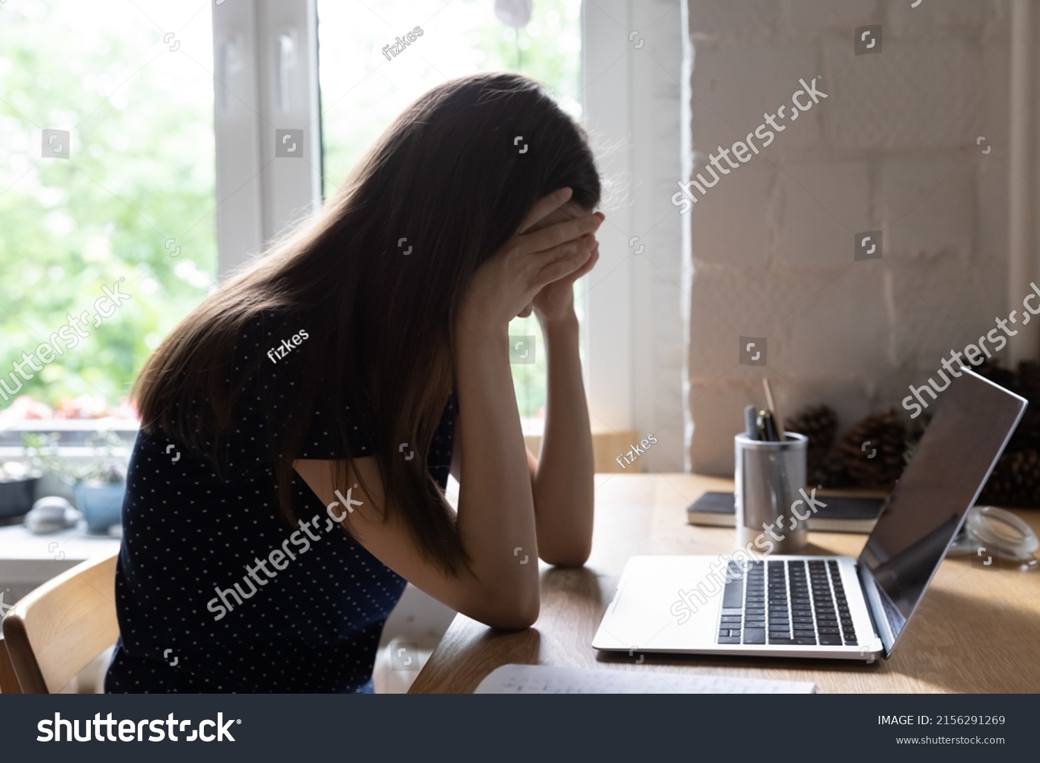 Exhausted upset young student girl sitting at table with laptop, leaning head on hand, suffering from headache, feeling depressed, unhappy, tired, getting bad news. Professional burnout concept #2156291269