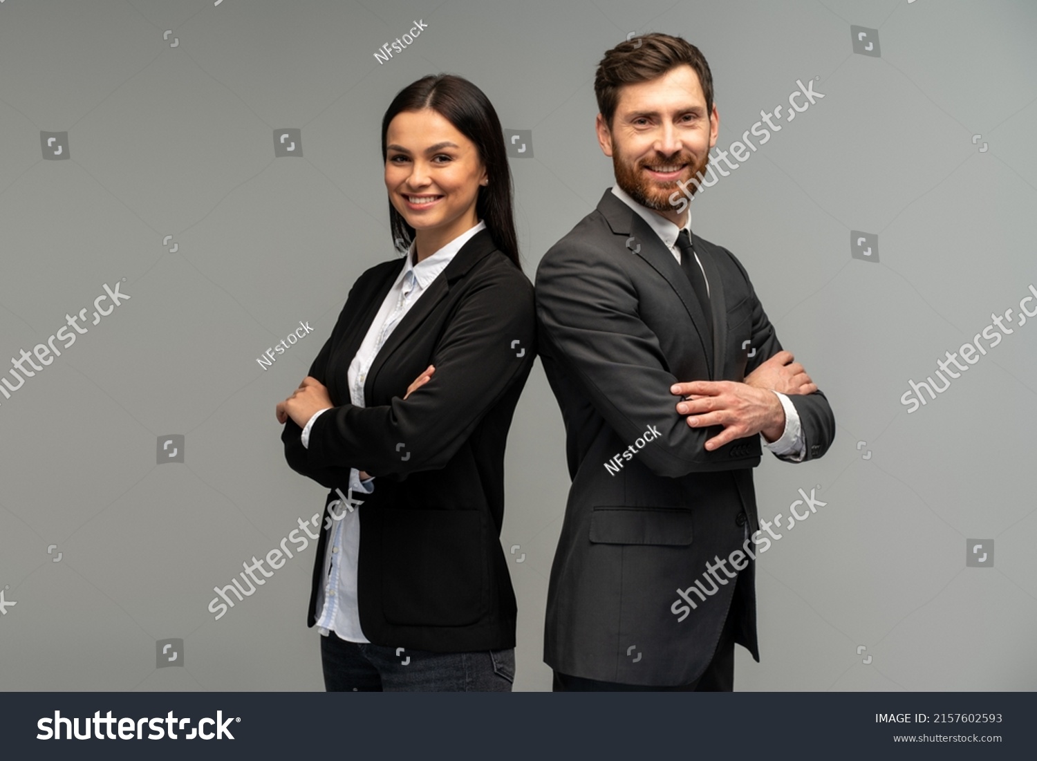 Concept of partnership in business. Young man and woman standing back-to-back with crossed hands against grey background  #2157602593