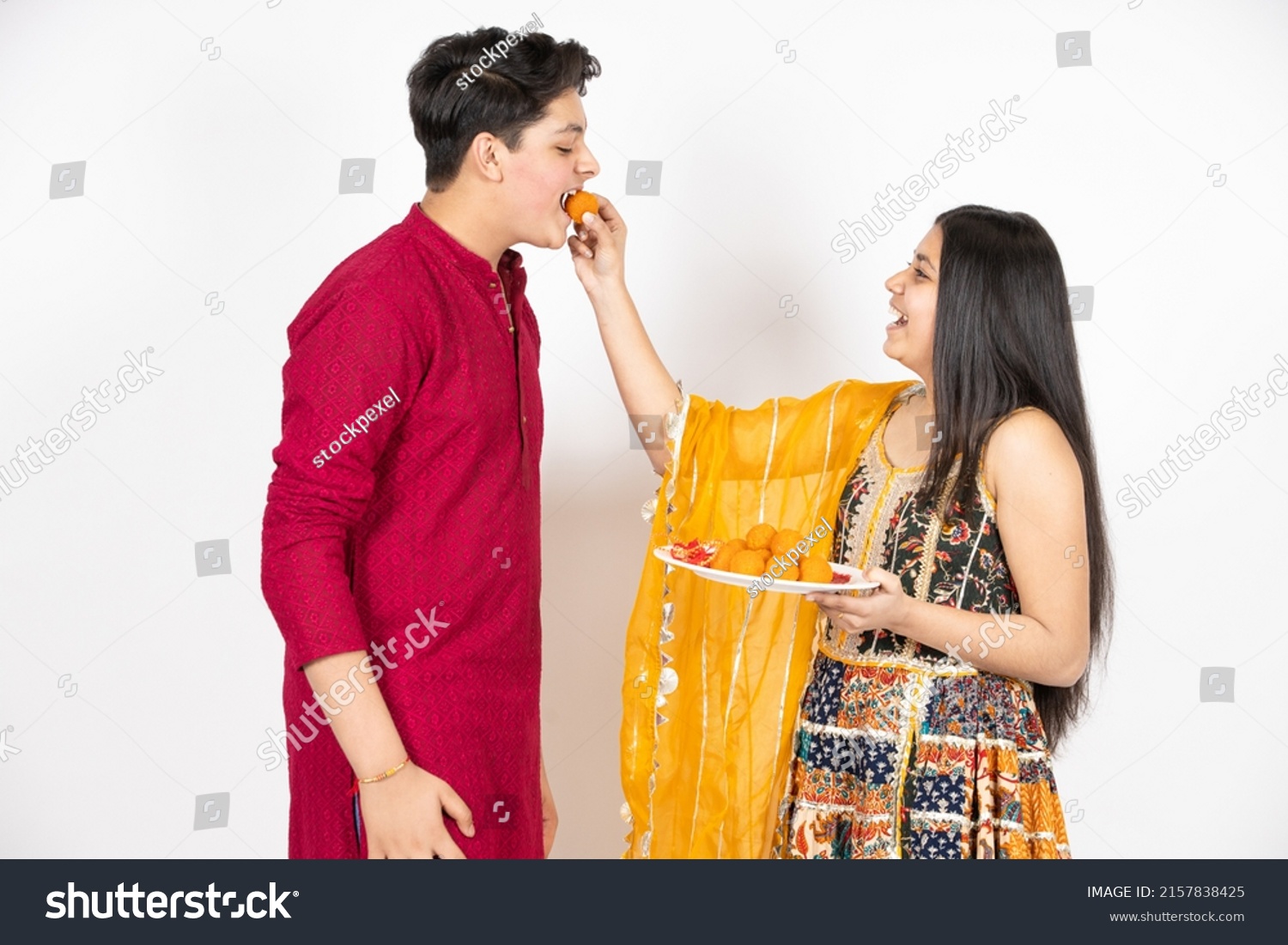 Young Indian brother and sister celebrating Diwali, rakshabandhan or bhai dooj festival isolated on white studio background. enjoying sweet laddo, #2157838425