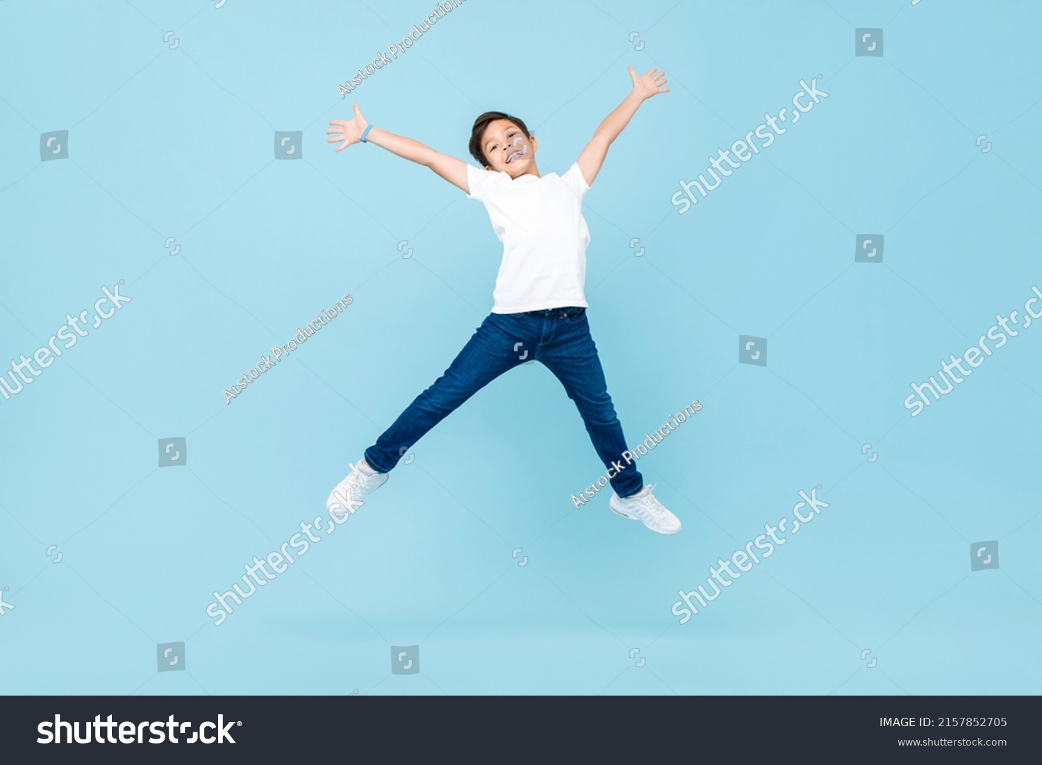 Energetic Asian mixed race boy jumping and raising hands up in isolated light blue color studio background #2157852705