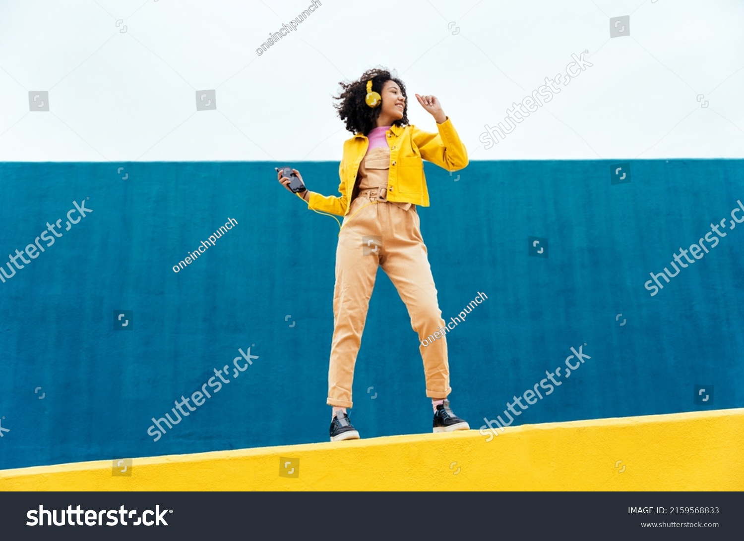 Young happy woman dancing and having fun outdoor. Teenager listening to music with smartphone and headphones in a yellow and blue modern urban area #2159568833