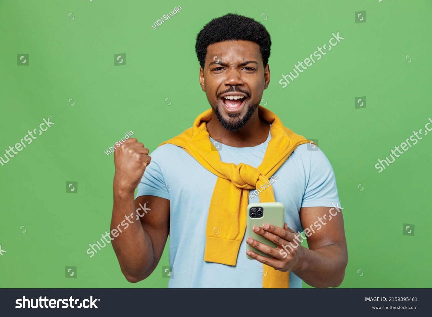 Young excited man of African American ethnicity 20s wear blue t-shirt hold in hand use mobile cell phone do winner gesture isolated on plain green background studio portrait. People lifestyle concept #2159895461