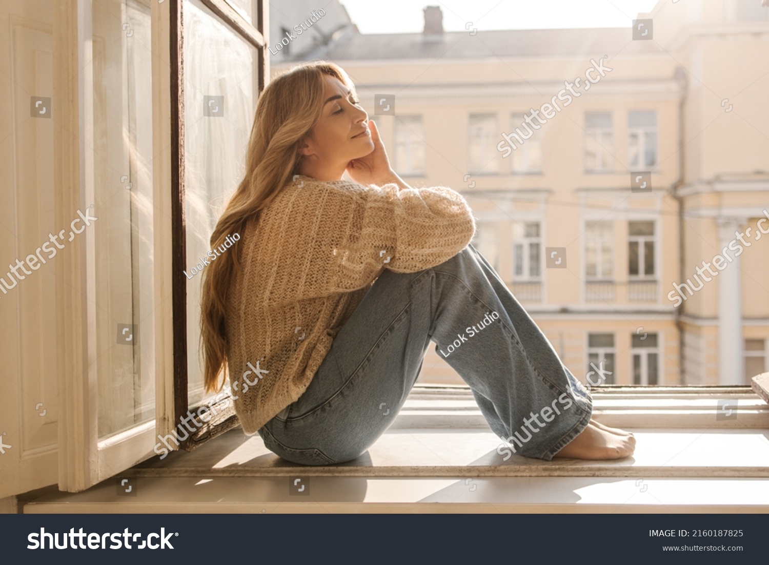 Cute caucasian adult lady with her eyes closed is basking in sun sitting on windowsill on warm day. Long-haired blonde wears casual clothes. Concept of enjoying moment #2160187825