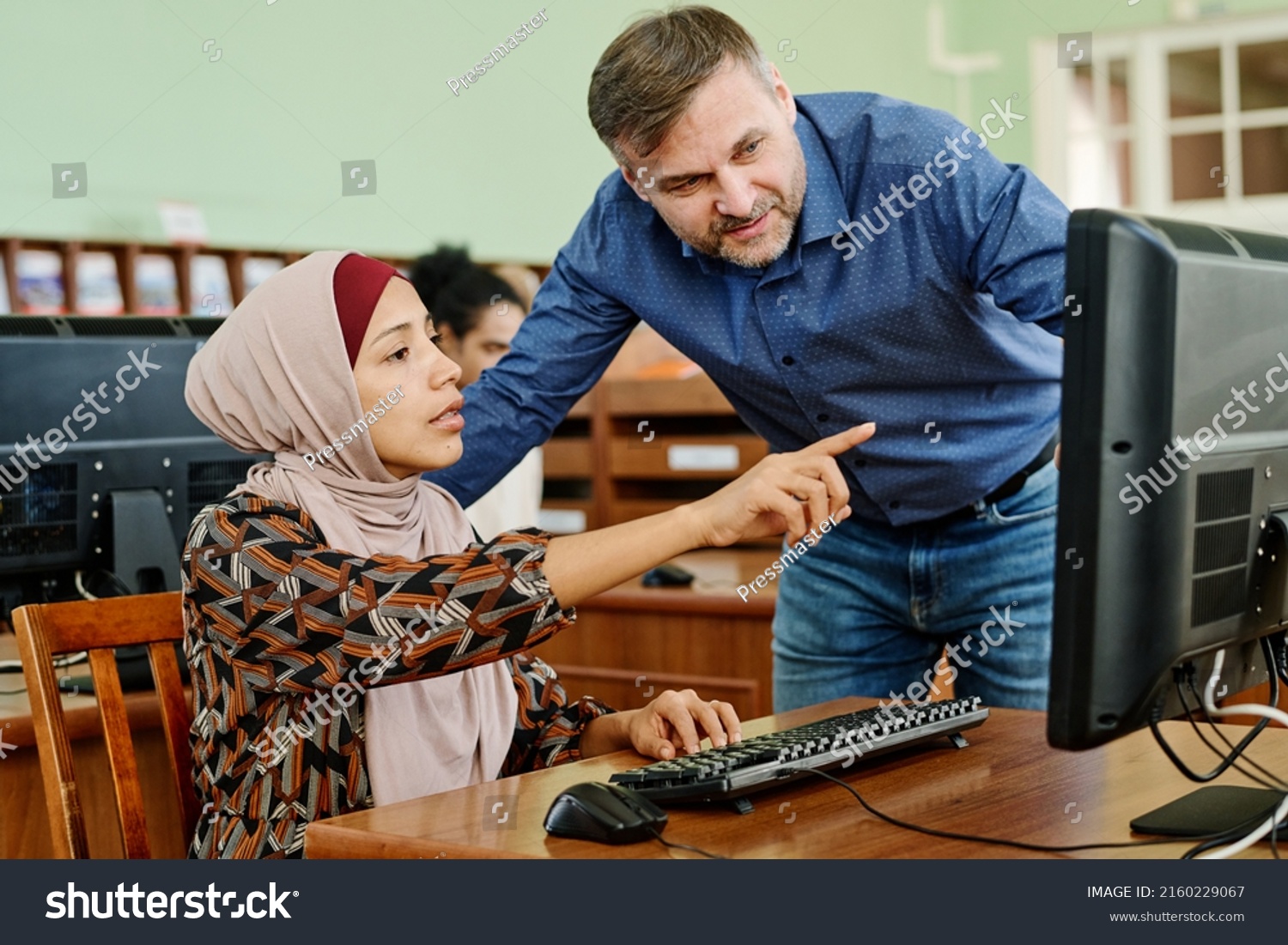 Young Muslim woman wearing hijab doing difficult task on desktop computer asking teacher to help with it #2160229067