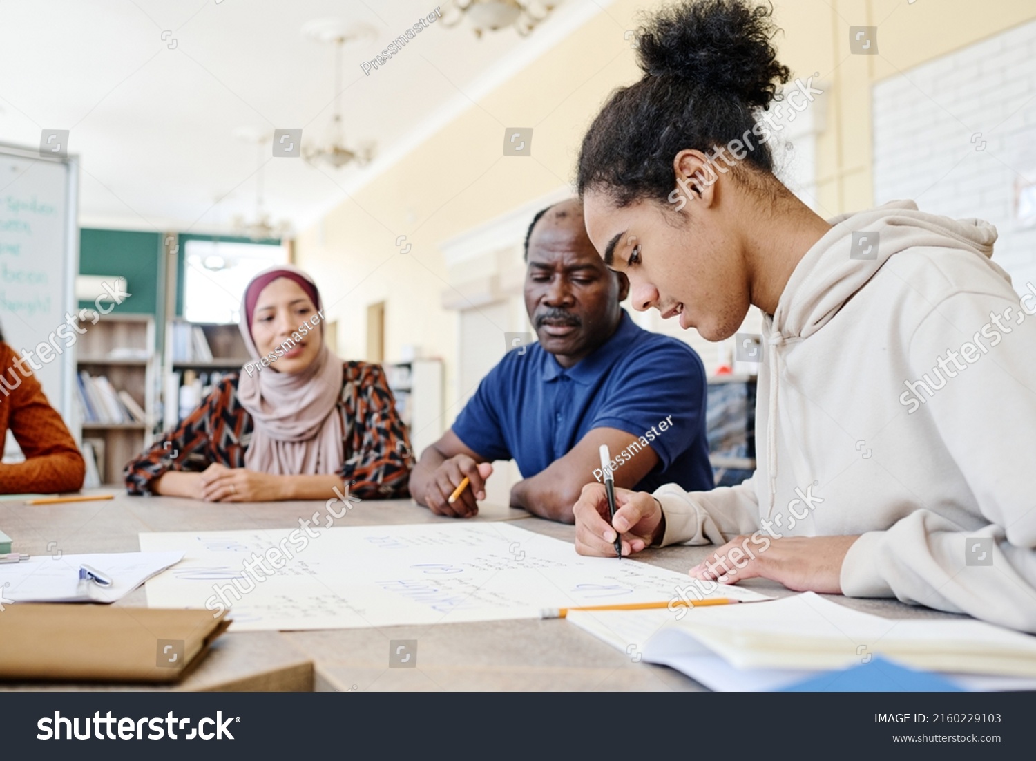 Young Black immigrant sitting at table writing something on handmade educational English language poster during lesson #2160229103