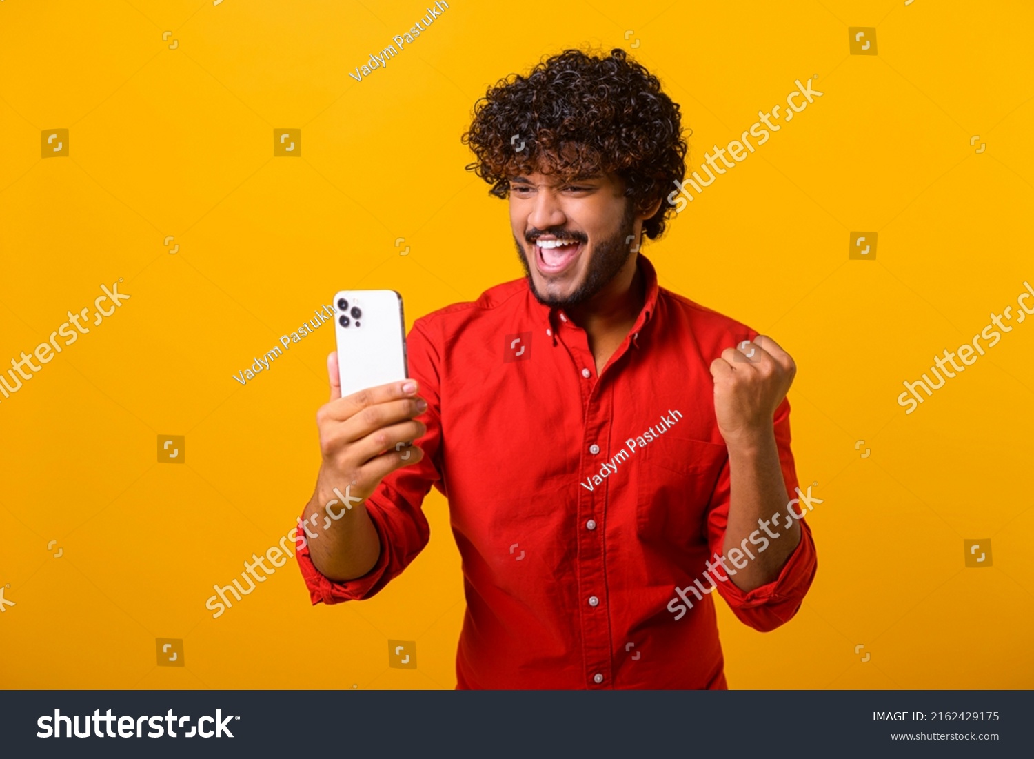 Happy satisfied man with beard holding smartphone and smiling making yes gesture, celebrating online lottery or giveaway victory. Indoor studio shot isolated on orange background #2162429175