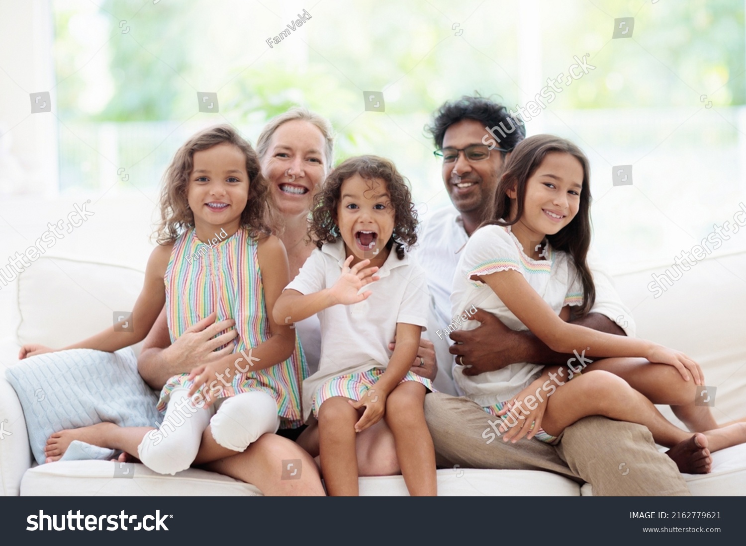 Happy family with kids at home. Parents and children sitting on white couch in sunny living room. Mother, father, son and daughter play and laugh. Young beautiful interracial couple in new house. #2162779621