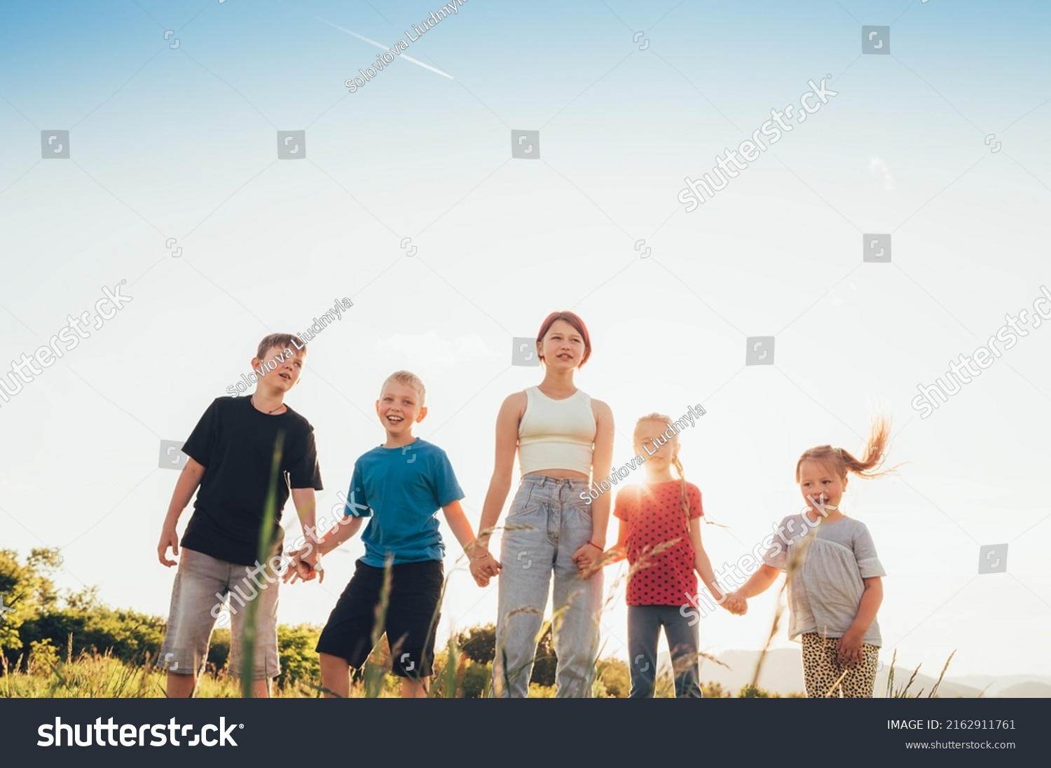Five kids brothers and sisters teenagers and little kids walking on the green grass meadow with evening sunset background light holding hands in hands and smiling. Happy and careless childhood concept #2162911761