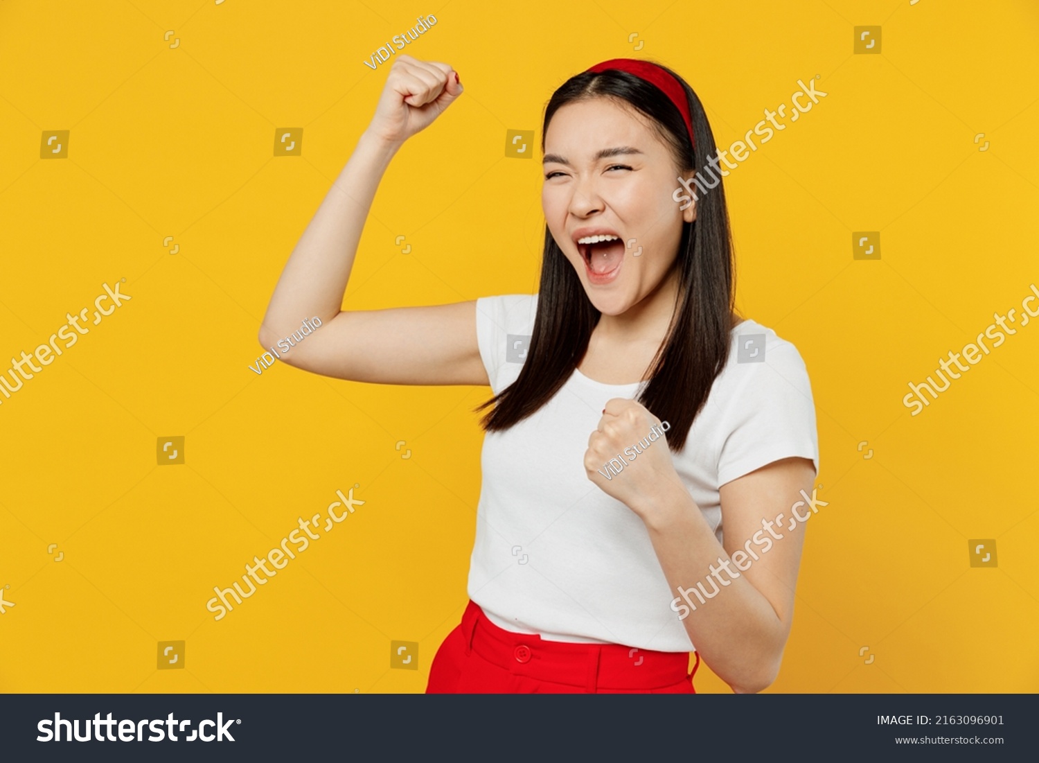 Happy fun excited jubilant young woman of Asian ethnicity 20s years old wears white t-shirt doing winner gesture celebrate clenching fists say yes isolated on plain yellow background studio portrait #2163096901