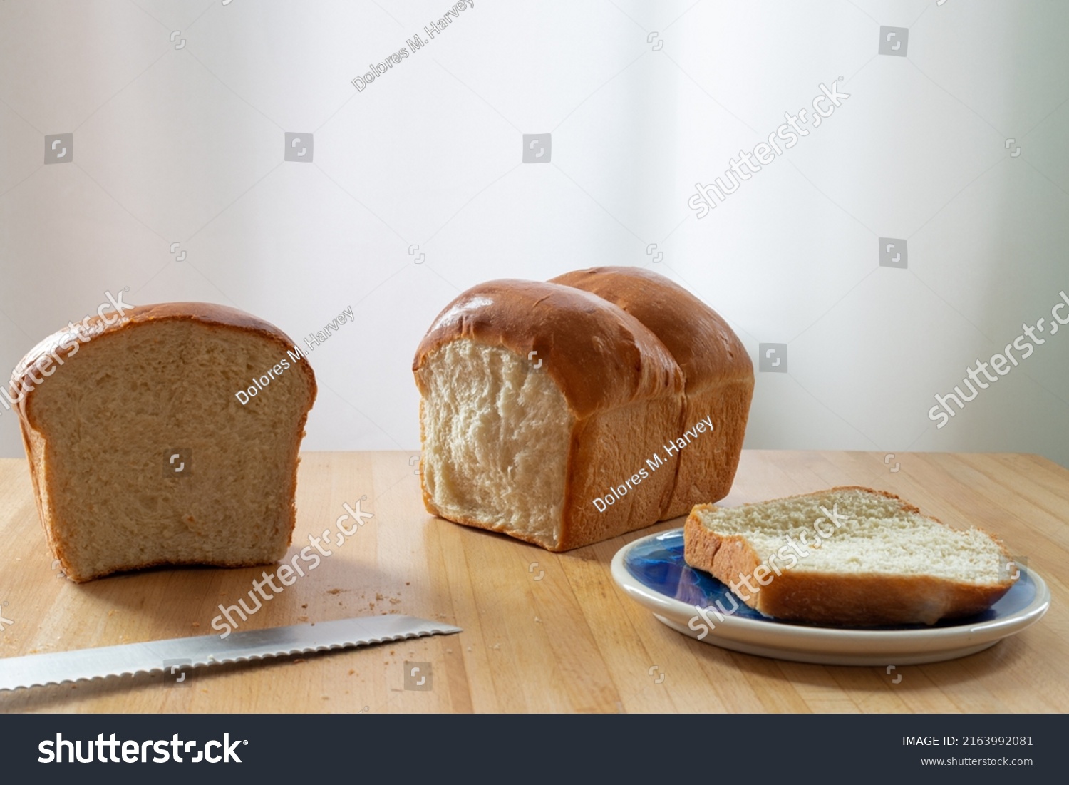 A single loaf of fresh white crusty bread on a wooden cutting board on a kitchen table. The warm crisp bun has melted butter over the crisp loaf buns. Side plates and a knife are next to the loaf.  #2163992081