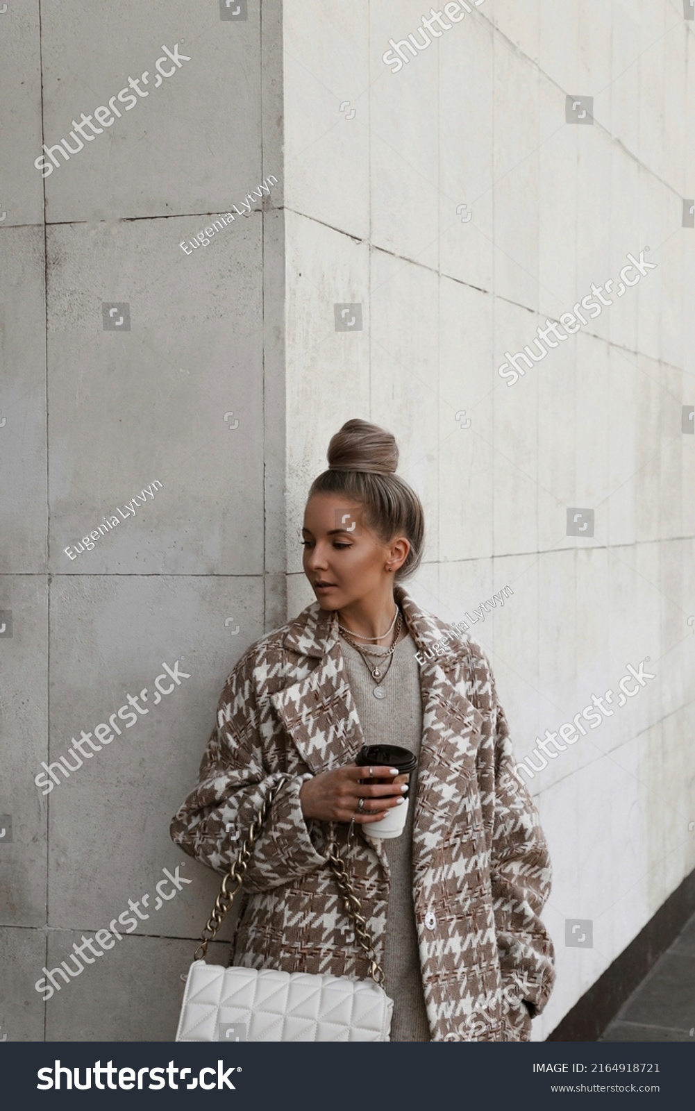 portrait of beautiful blonde girl dressed in oversize beige coat with print, knitted dress, white bag in hands, hair gathered in bun, stylish trendy fashion outlook, lifestyle model #2164918721