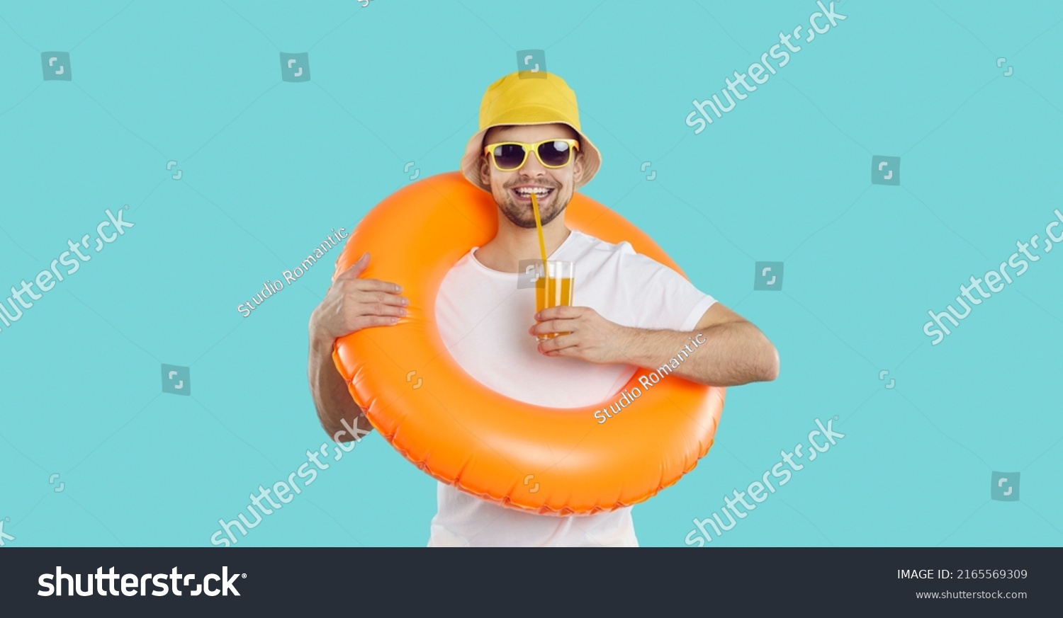 Happy young man with inflatable swimming circle and with glass of juice enjoys summer vacation. Cheerful funny guy in summer clothes drinks juice on light blue background. Summer relaxation concept. #2165569309