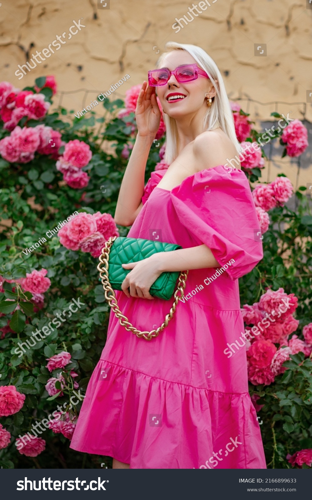 Happy smiling fashionable woman wearing trendy summer fuchsia color dress, pink sunglasses, holding stylish green faux leather bag, posing near blooming roses #2166899633
