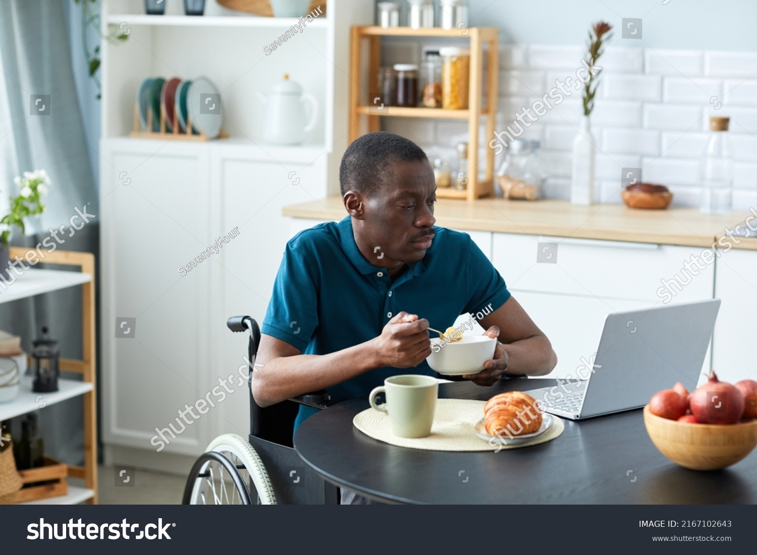 Portrait of black adult man with disability eating breakfast at table in kitchen and watching videos, copy space #2167102643