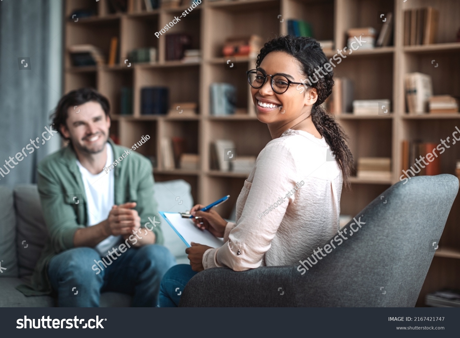 Happy millennial black woman psychologist in glasses look at camera, consulting smiling european man in office clinic interior. Therapy, mental problems, psychologist support and professional help #2167421747