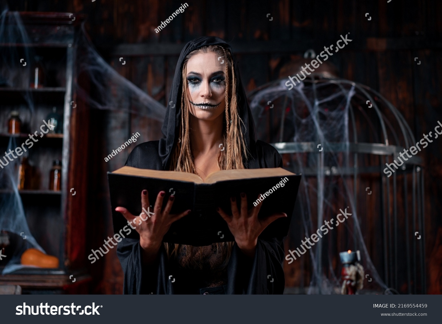 Halloween concept. Witch dressed black hood with dreadlocks standing dark dungeon room use magic book for conjuring magic spell. Female necromancer wizard gothic interior looking camera medium shoot #2169554459