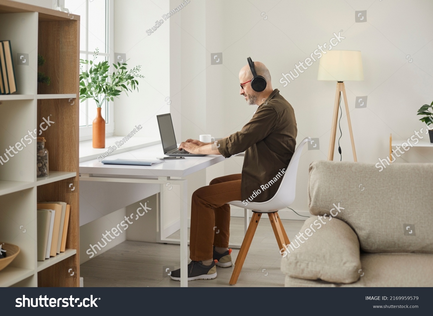 Smiling adult man listening to music on headphones while working on laptop at home. Side view of middle-aged man listening to his favorite music, searching information on Internet or making video call #2169959579
