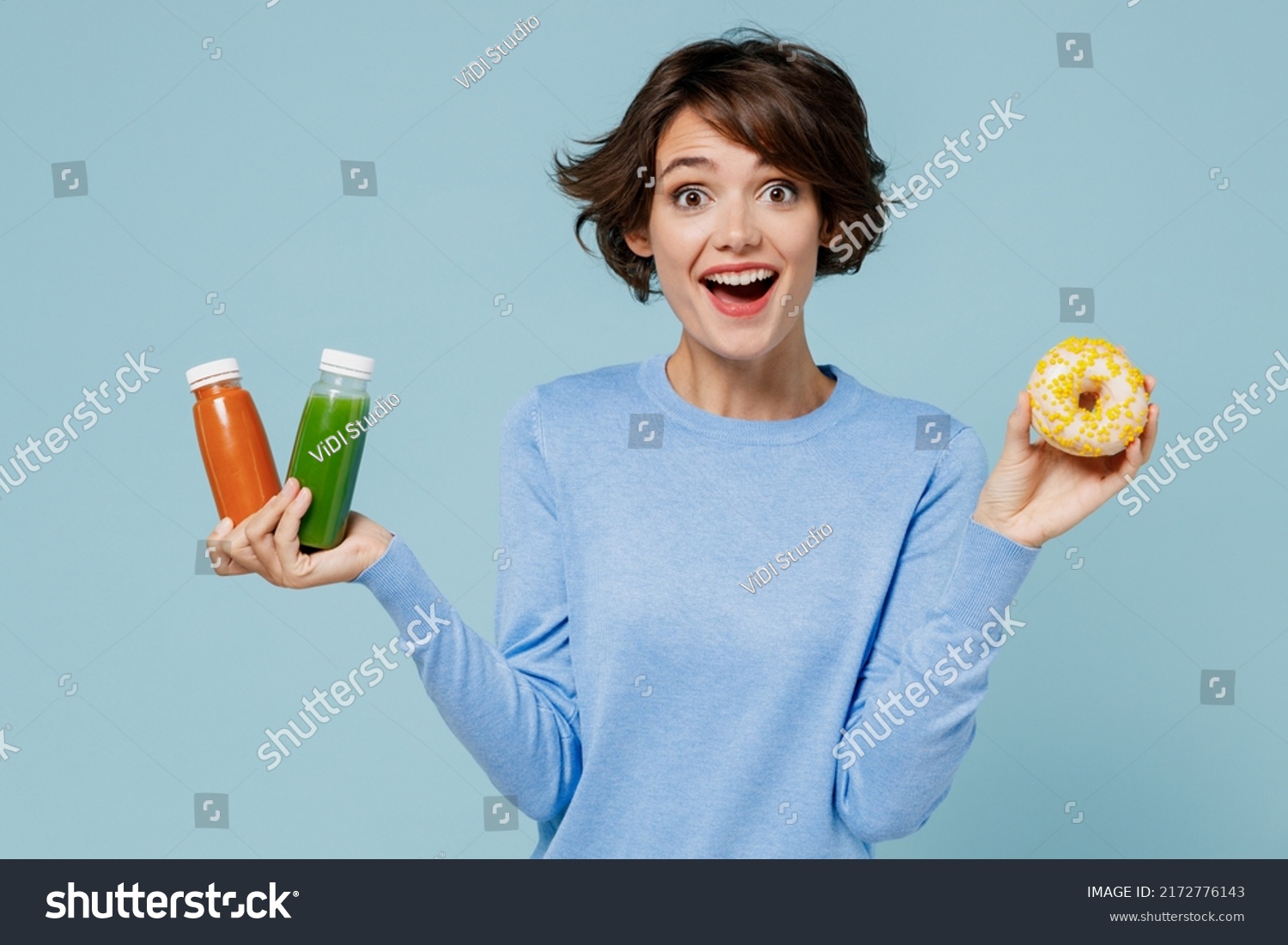 Young surprised woman in casual sweater hold pressed juice green orange vegetable smoothie as detox diet put arm on belly isolated on plain pastel light blue background. People lifestyle food concept #2172776143