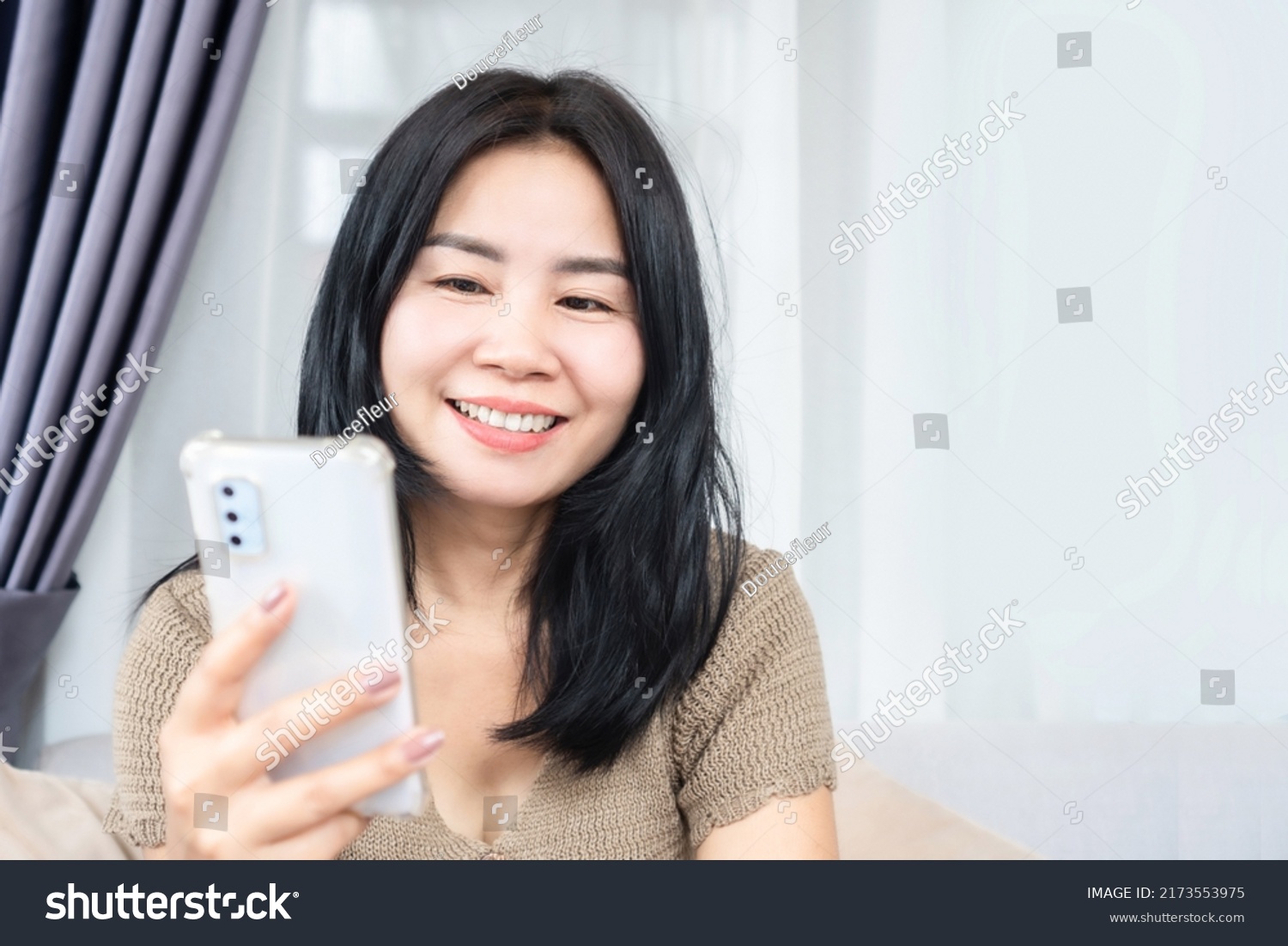smiling Asian female sitting on sofa using mobile phone enjoin surfing the internet on the weekend #2173553975