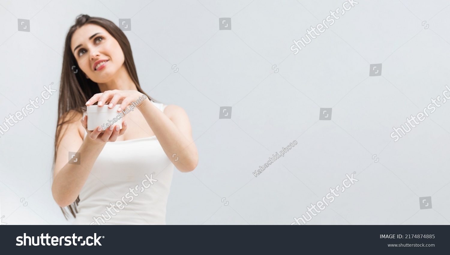 Positive woman opens jar of nourishing cream on light background in studio #2174874885
