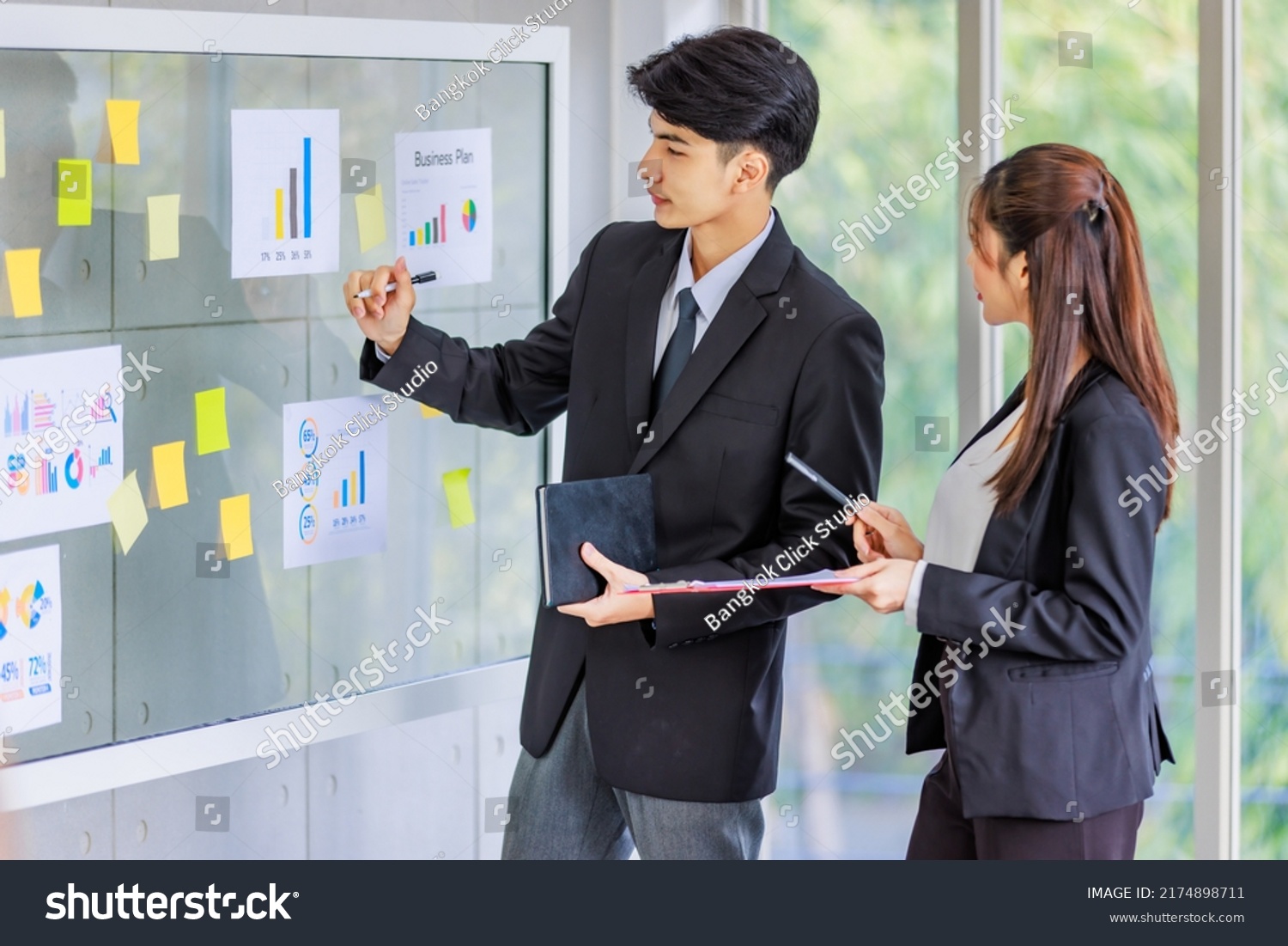 Asian young male professional successful businessman manager pointing paperwork company target graph chart diagram document on glass board explaining to female secretary employee staff in formal suit. #2174898711