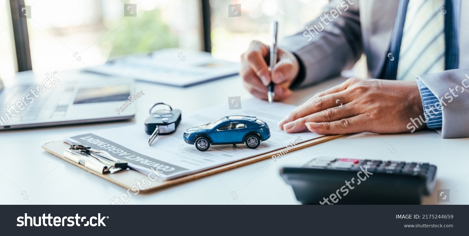 Man signing car insurance document or lease paper. Writing signature on contract or agreement. Buying or selling new or used vehicle. Car keys on table. Warranty or guarantee. Customer or salesman. #2175244659