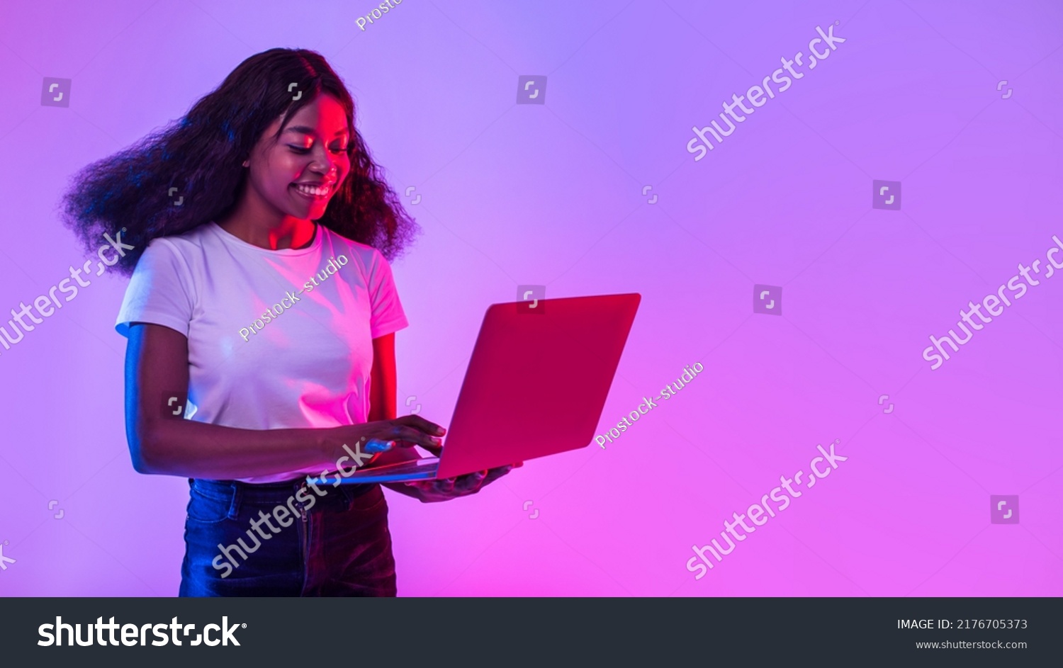 Portrait of cheery young black woman using laptop pc, studying or working remotely, having online conference in neon light, banner with empty space. African American lady with computer browsing web #2176705373