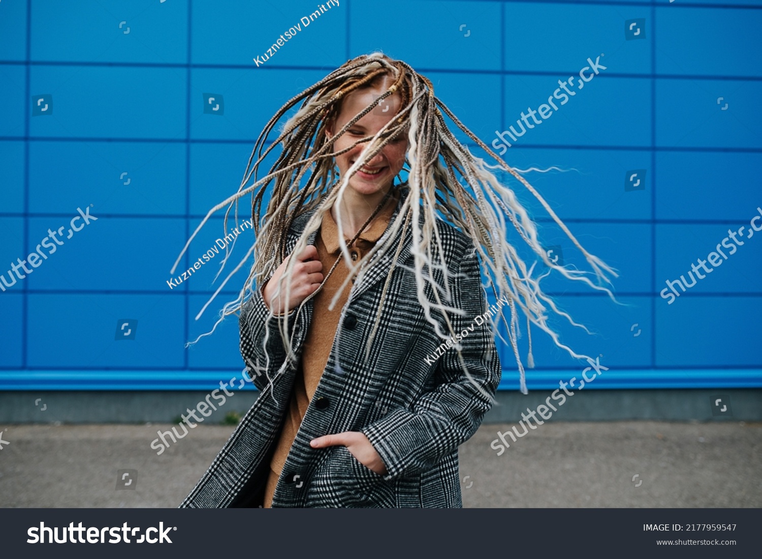 Shaking dreads lighthearted teenage girl in front of a blue panel wall covering. She is wearing a grey checkered jacket. Portrait. #2177959547