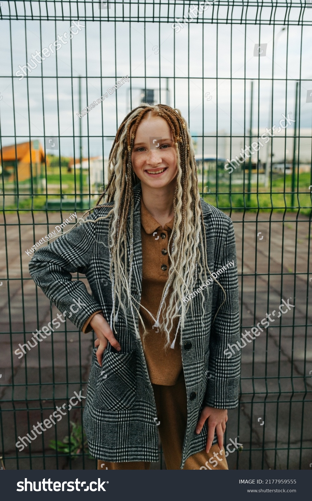 Lighthearted teenage girl with dreads standing in front of a metal fence. She is wearing a jacket. Portrait. #2177959555