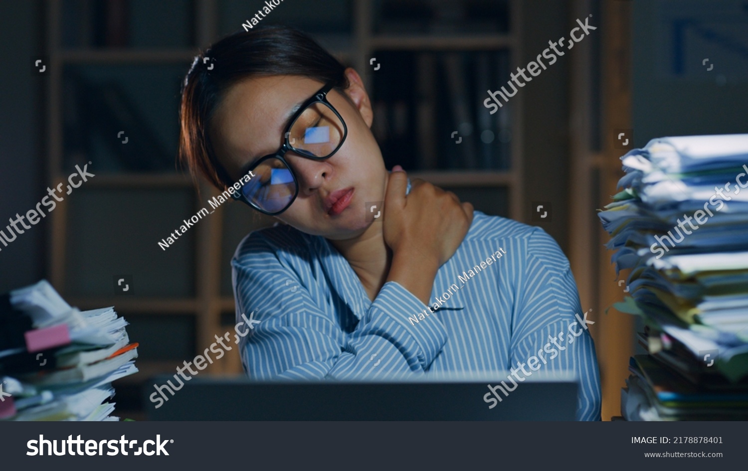 Tired Asian office employee massaging neck and shoulder muscles fatigued from using laptop computer overtime at night in office, office syndrome #2178878401