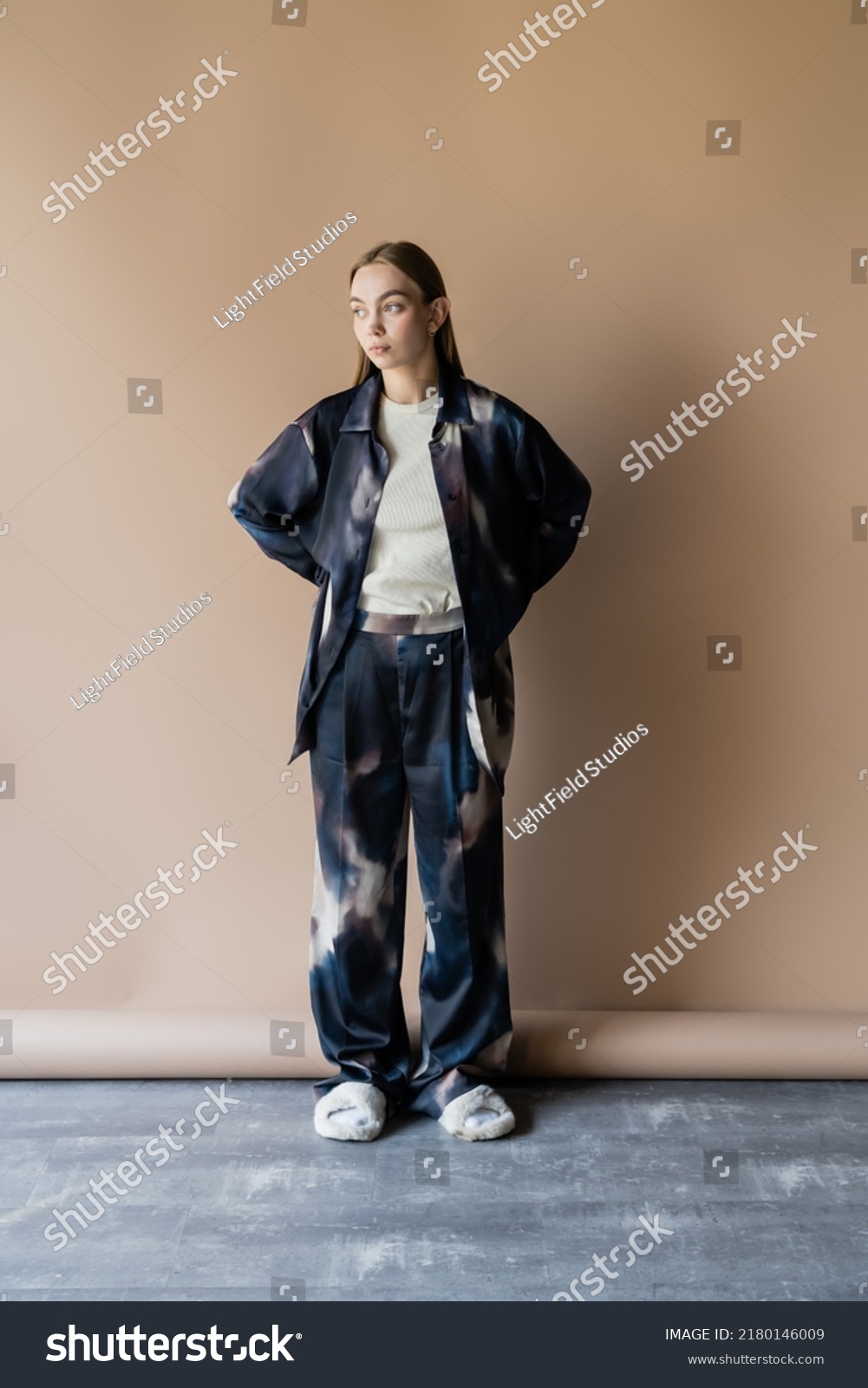 full length of woman in oversize clothes and soft slippers posing with hands behind back on beige background #2180146009
