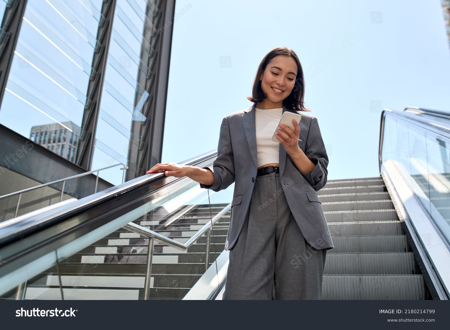 Smiling young Asian business woman wearing suit standing on urban escalator using applications on cell phone, reading news on smartphone, fast connection, checking mobile apps outdoors. #2180214799