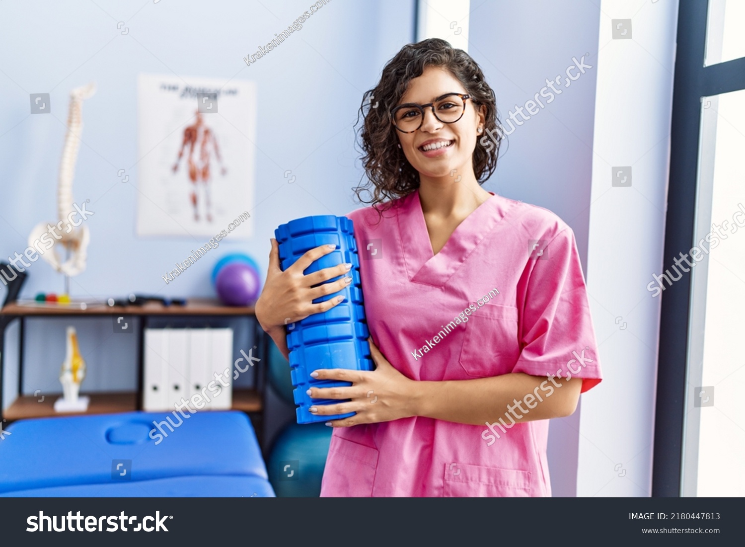 Young latin woman wearing physiotherapist uniform holding foam roller at clinic #2180447813