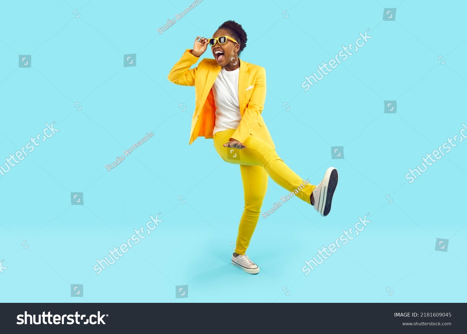 Cheerful young woman having fun, dancing and laughing isolated on light blue background. African American young woman in yellow suit and sunglasses is walking with funny expression on her face. #2181609045