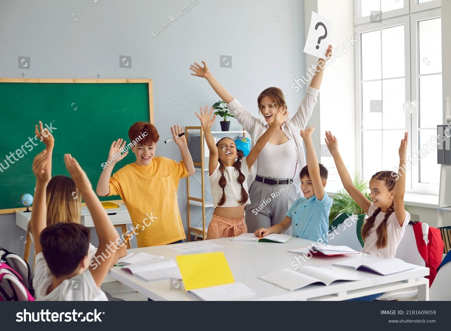 Happy teacher and children having fun in class. Cheerful woman and joyful students play games in the classroom, write in notebooks, answer questions and solve problems together. Back to school concept #2181609059