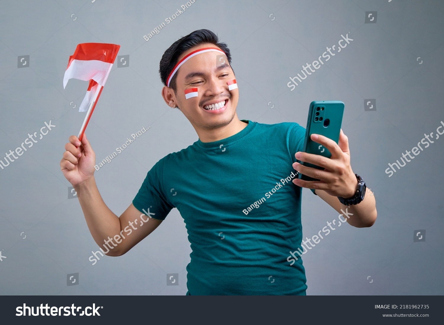 Excited young Asian man holding mobile phone and indonesian flag while celebrating indonesia independence day isolated on grey background. indonesia independence day celebration concept #2181962735
