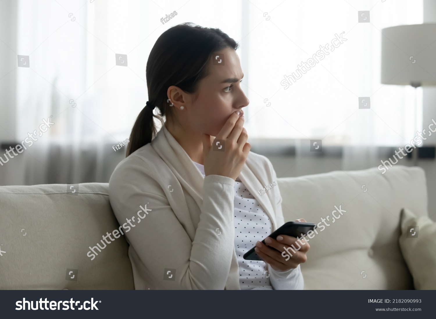 Pensive stressed young Caucasian woman sit on sofa at home using cellphone look in distance pondering of problem. Anxious millennial girl feel distressed frustrated with message or text on smartphone. #2182090993