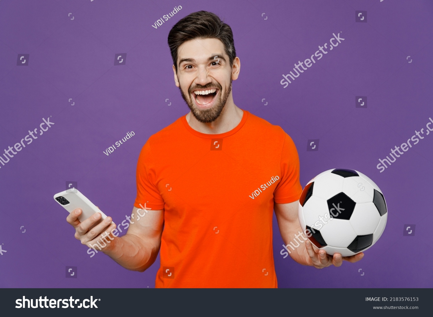 Young fun fan happy cheerful man he wears orange t-shirt cheer up support football sport team hold in hand soccer ball watch tv live stream use mobile cell phone isolated on plain purple background #2183576153