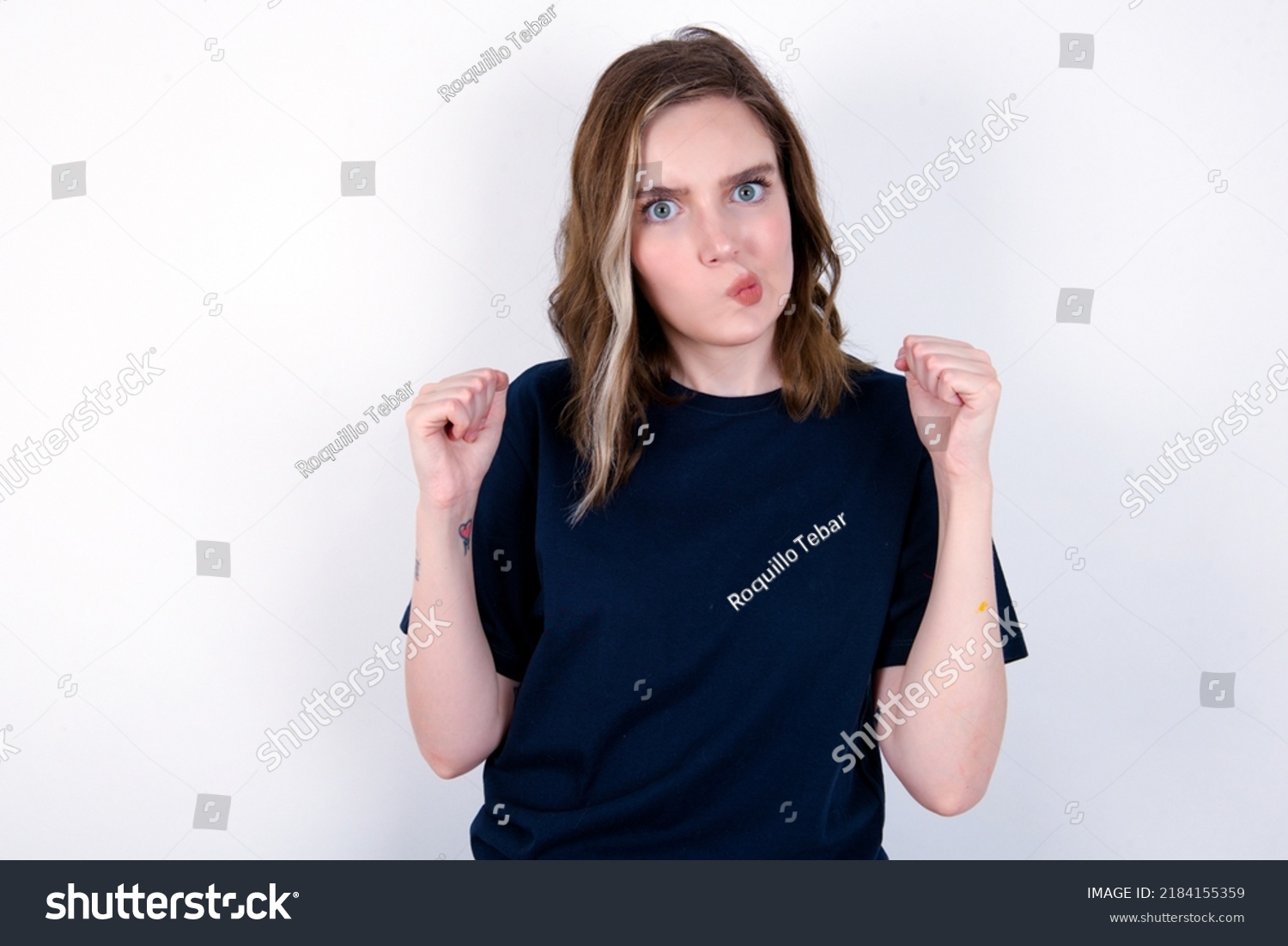 Irritated young caucasian woman wearing black T-shirt over white background blows cheeks with anger and raises clenched fists expresses rage and aggressive emotions. Furious model #2184155359