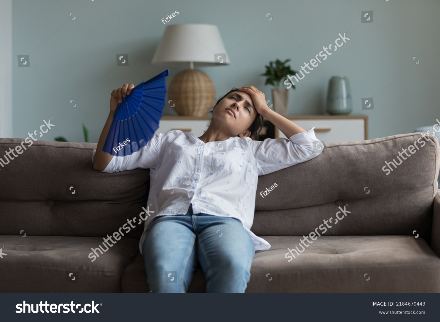 Exhausted young Indian girl feeling hot, tired, sick, resting on couch at home, waving paper handheld fan for cooking, suffering from heat attack, hypoxia, headache, touching head #2184679443
