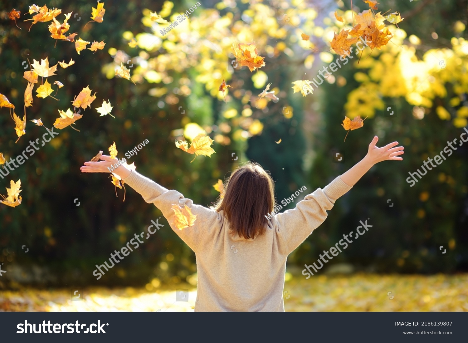 Young woman is having fun while walking through the forest on a sunny autumn day. Girl plays with maple leaves and throws them up. Fallen leaves rustle. #2186139807