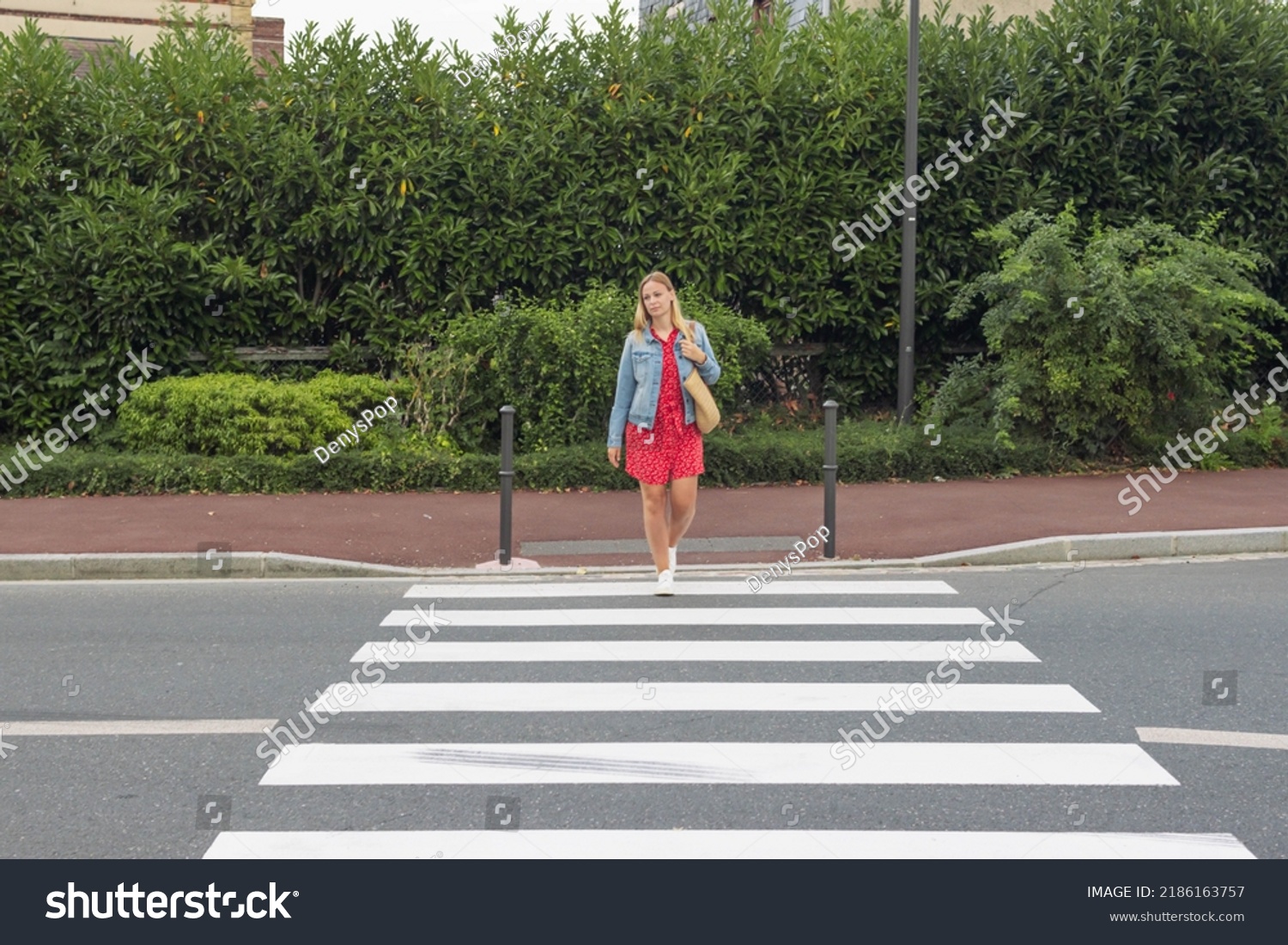 A girl with fair hair in a red dress and a denim jacket with a bag on her shoulder crosses the road at a pedestrian crossing. The concept of traffic rules #2186163757