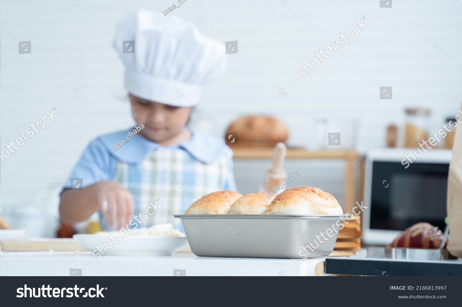 Selective focus on finished fresh homemade bread in loaf pan with little kid girl wear chef hat and apron have fun holding rolling pin to knead dough bread in kitchen at home. Child education concept #2186813997