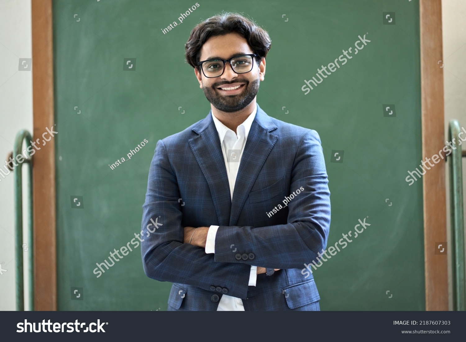 Smiling young indian business man professional manager wearing suit looking at camera standing arms crossed in office. Arab teacher or professor posing for portrait at work desk in front of #2187607303
