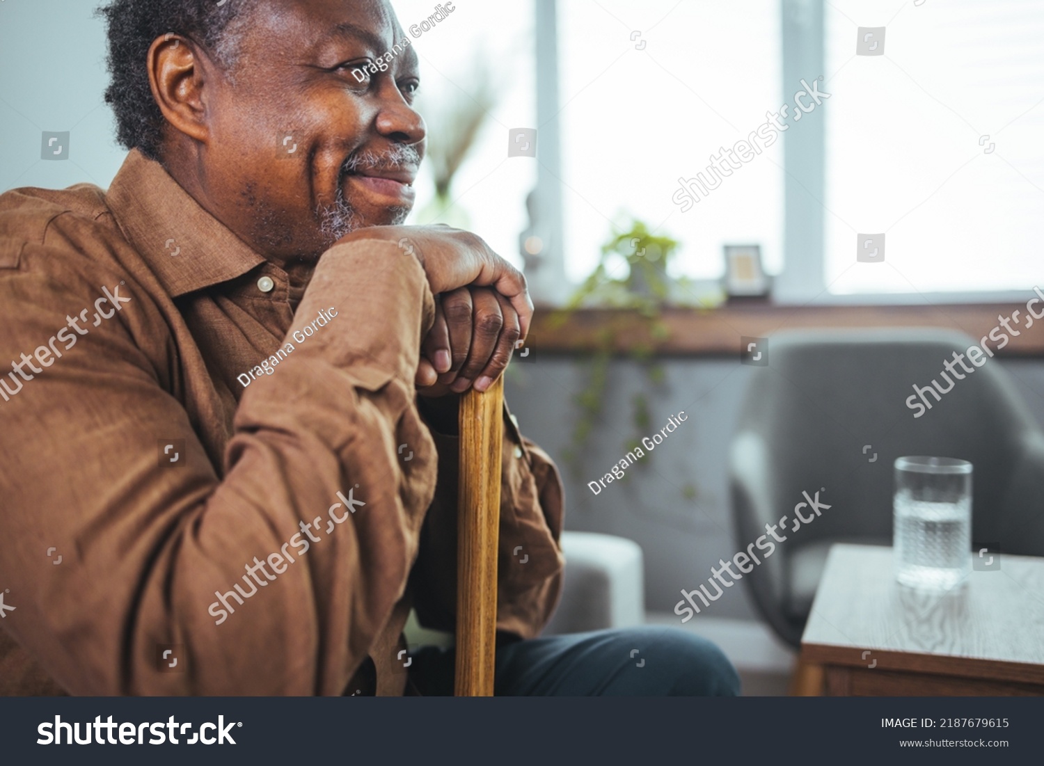 Shot of a senior man looking thoughtfully out of a window at home. Positive African American senior grandfather with grey hair and beard sitting at home. Elderly man sitting alone at home #2187679615